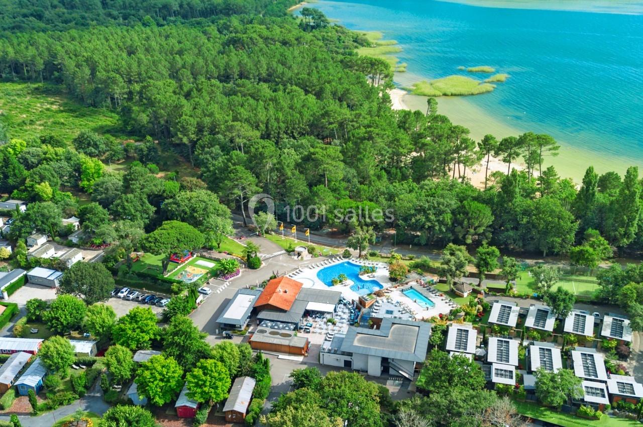 Vue aérienne d'un camping entouré de forêt, avec piscine, mobil-homes et accès à un lac bordé de végétation.