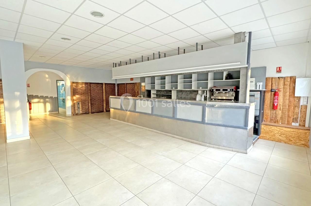 Intérieur d'un espace vide avec un comptoir de bar en métal, murs en briques et carrelage clair au sol.