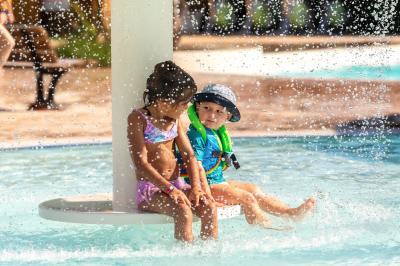 Un enfant avec des brassards orange glisse sur un toboggan rouge dans une piscine extérieure ensoleillée.