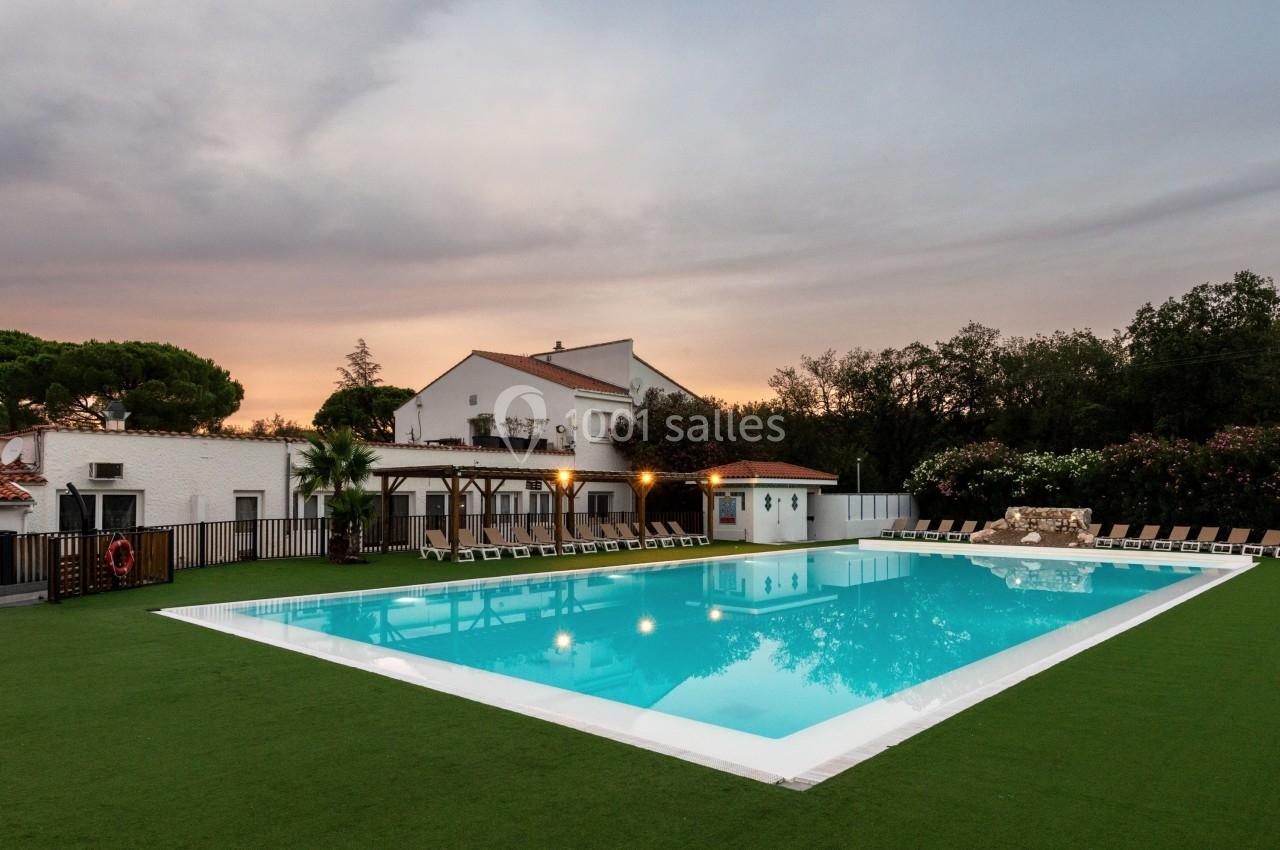 Piscine extérieure entourée de chaises longues, avec une maison blanche et un ciel nuageux en arrière-plan.