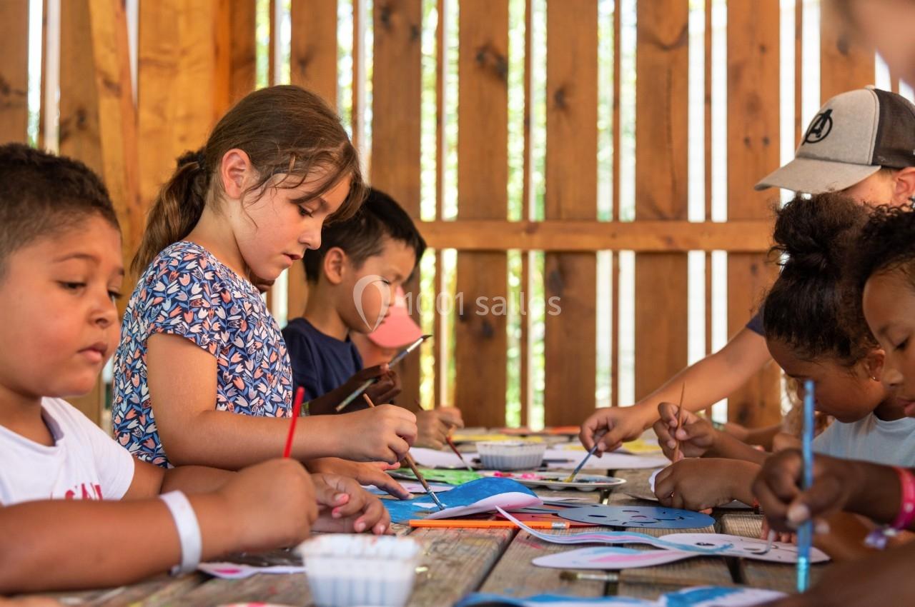 Des enfants peignent et dessinent ensemble sur une table en bois dans un espace extérieur lumineux.