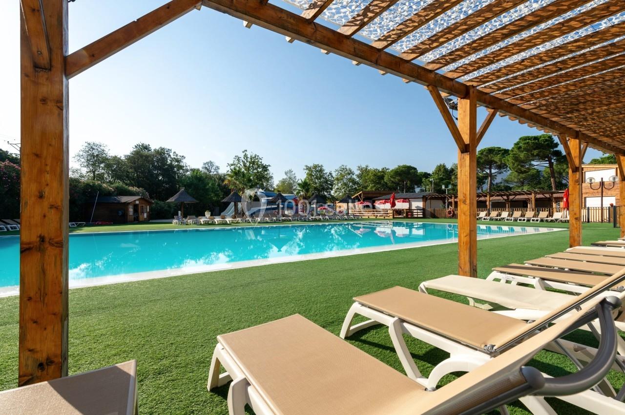 Piscine extérieure entourée de transats sous une pergola en bois, avec un ciel dégagé et des arbres en arrière-plan.