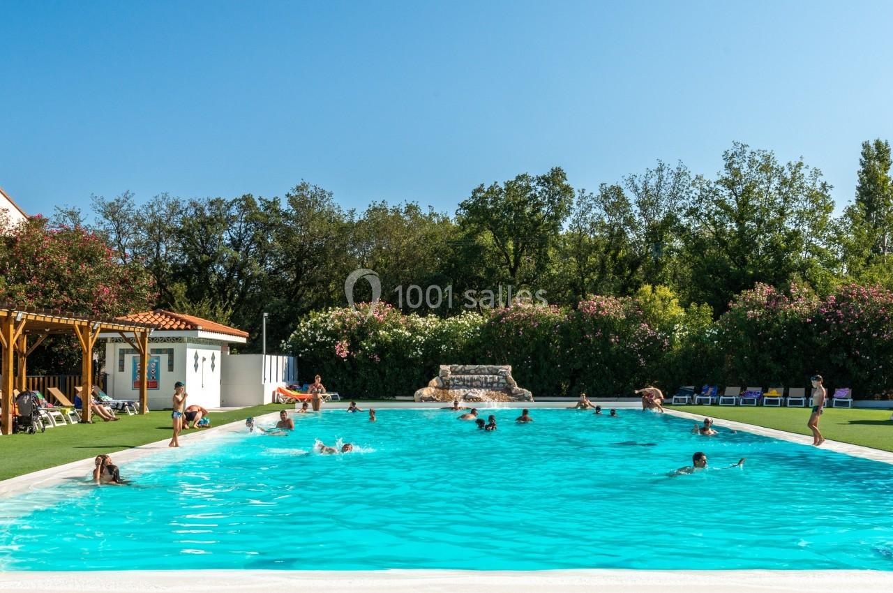 Piscine extérieure avec des baigneurs, entourée de verdure et de transats sous un ciel dégagé.