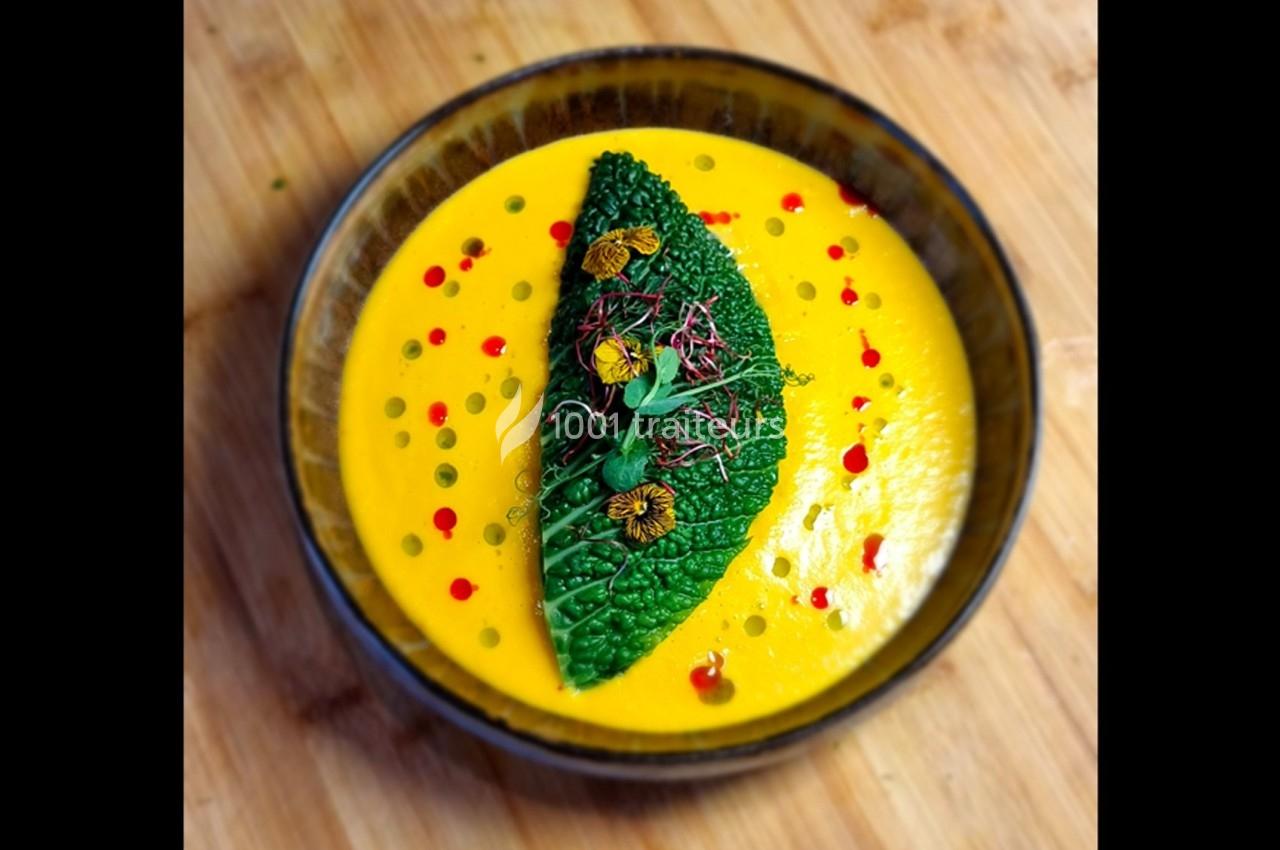 Bol de soupe jaune garnie d'une feuille de chou vert, de micro-pousses et de gouttes de sauce colorée.