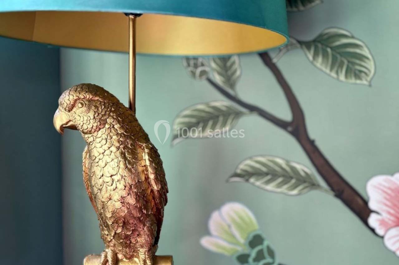 Lampe avec pied en forme de perroquet doré, posée devant un mur décoré de motifs floraux et feuillages.