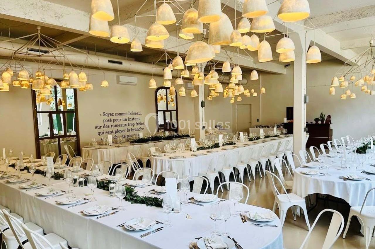 Salle de réception lumineuse avec de longues tables dressées, décorée de suspensions en forme de cloches.