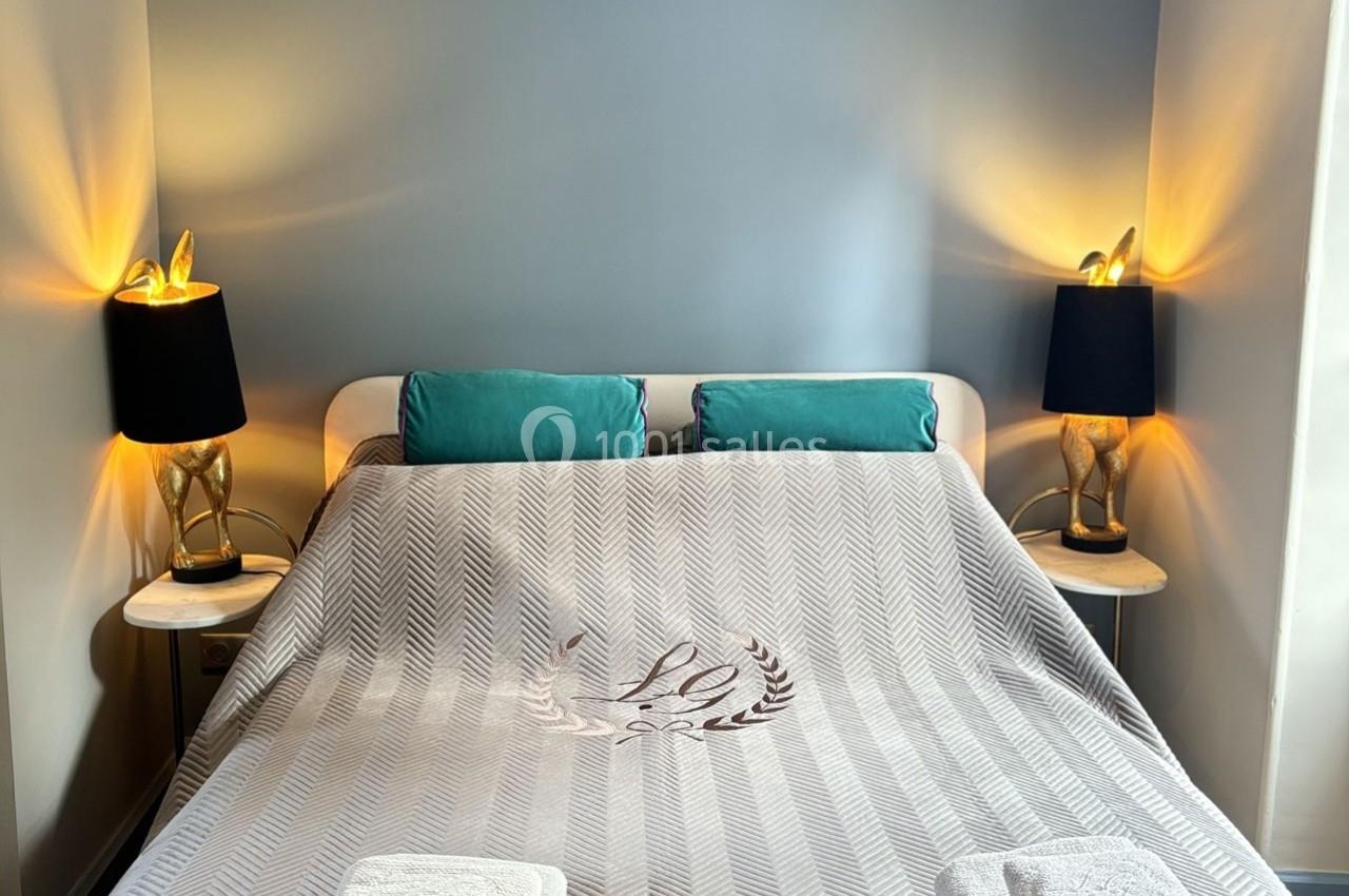 Lit double avec couverture grise, deux coussins turquoise et lampes de chevet allumées sur des tables en bois.