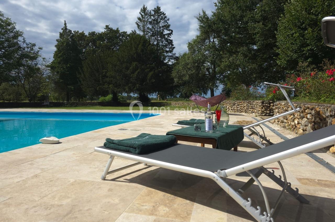 Deux transats et une table dressée près d'une piscine entourée de végétation et d'un mur en pierre.