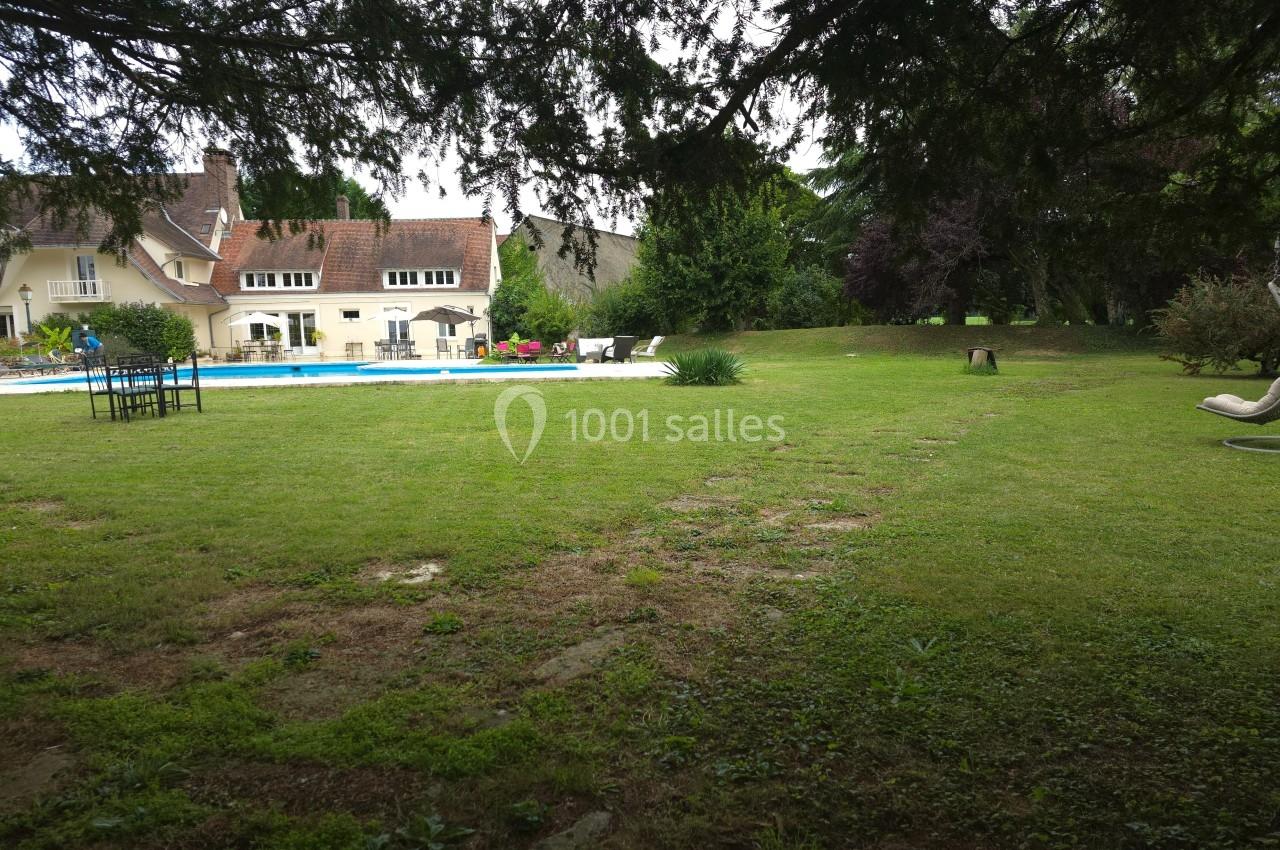 Jardin verdoyant avec pelouse, piscine, chaises longues et grande maison en arrière-plan sous un ciel couvert.