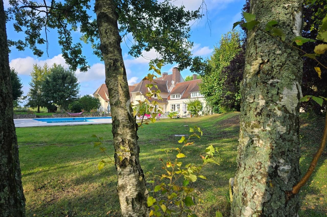Vue d'une maison entourée d'arbres, avec une pelouse et une piscine en arrière-plan sous un ciel bleu.