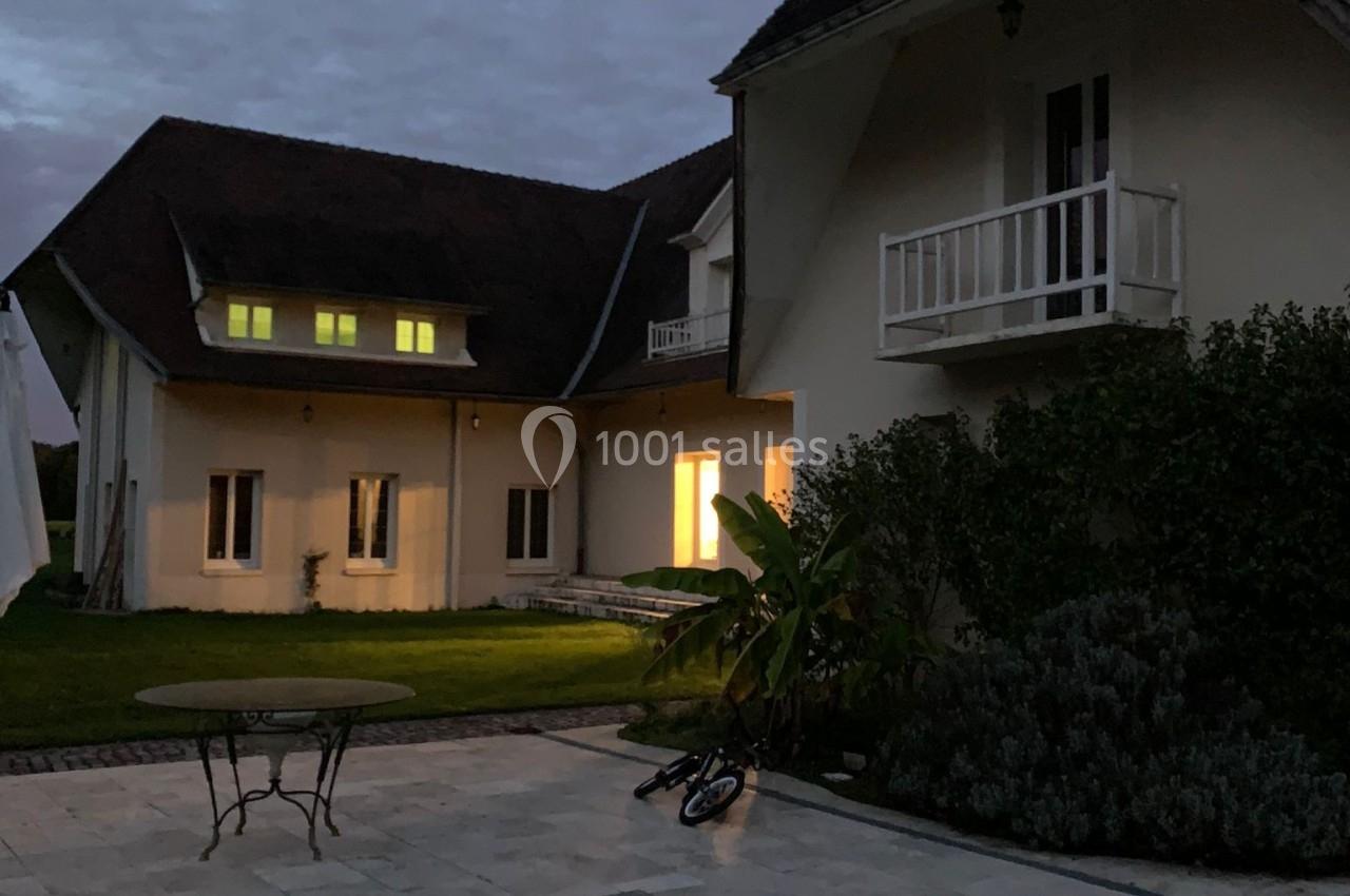 Grande maison avec toit en pente, éclairée de l'intérieur, vue depuis une terrasse en pierre au crépuscule.
