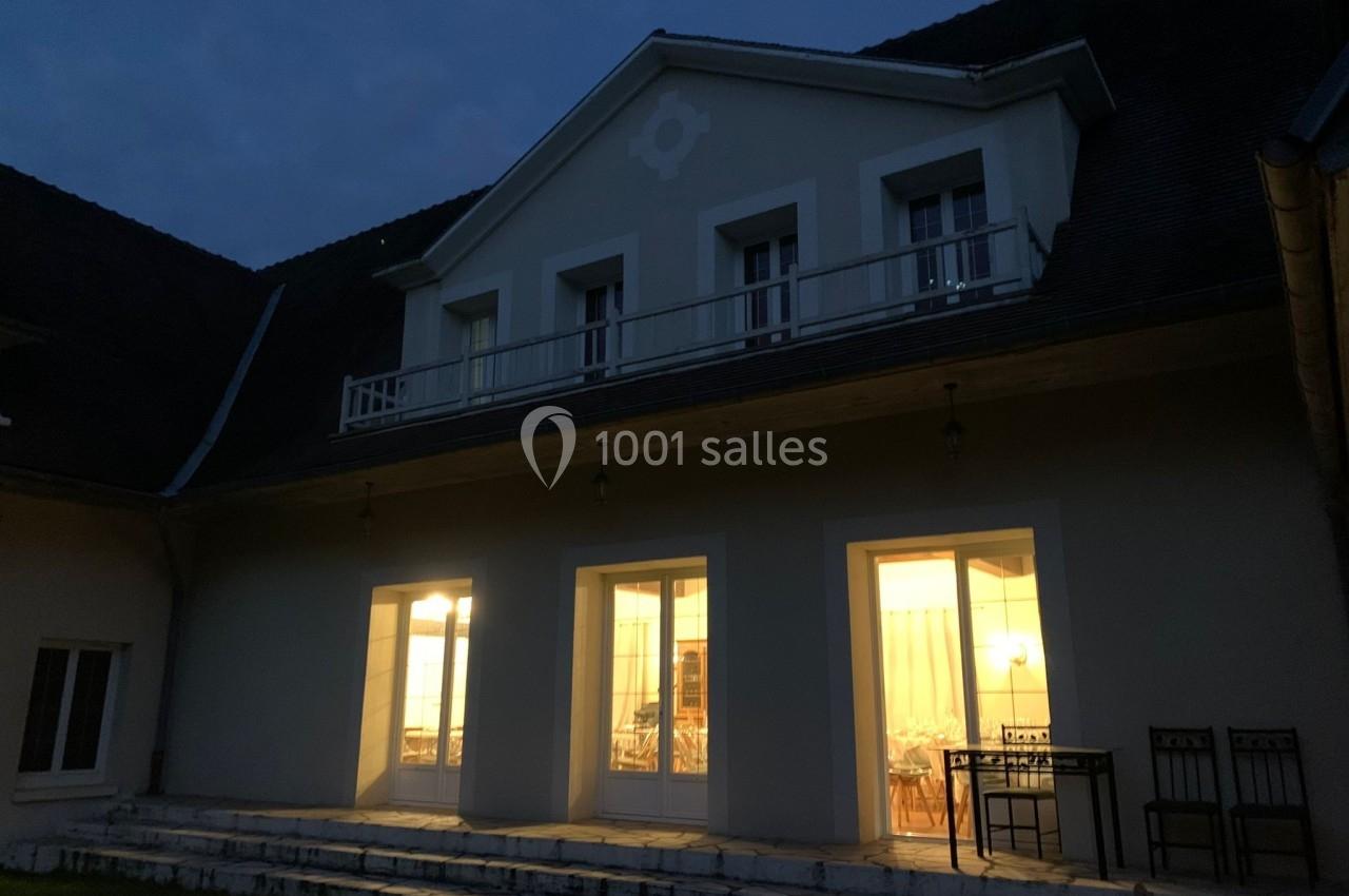 Façade d'une maison éclairée de nuit, avec des fenêtres et une terrasse visible au premier plan.