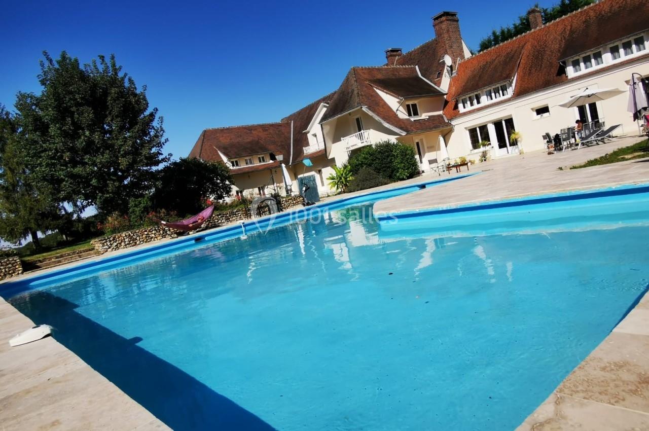 Piscine extérieure entourée de dalles en pierre, avec une grande maison traditionnelle et un ciel bleu en arrière-plan.