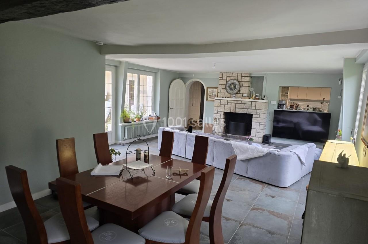 Salle à manger avec table en bois, chaises rembourrées, salon avec canapé blanc, cheminée en pierre et télévision.