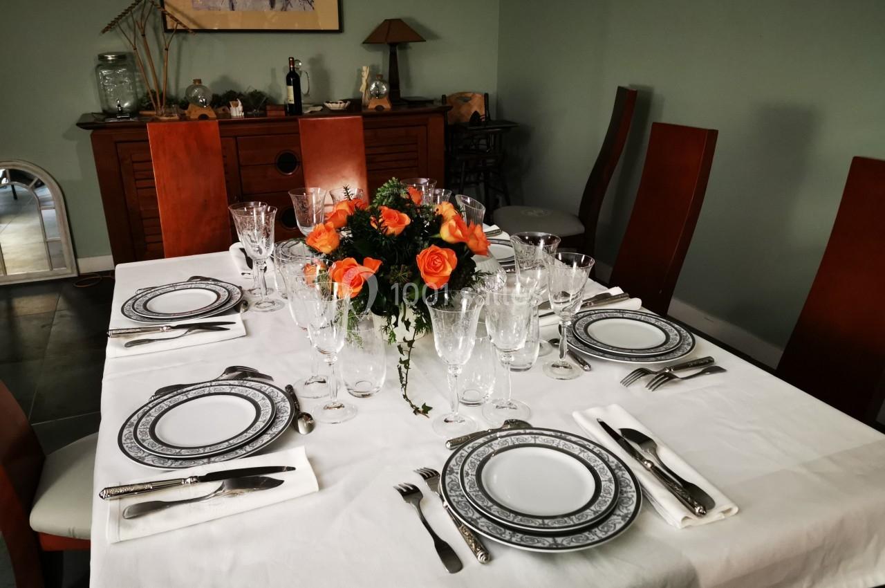 Table dressée avec une nappe blanche, vaisselle élégante, couverts disposés et un bouquet de roses orange au centre.