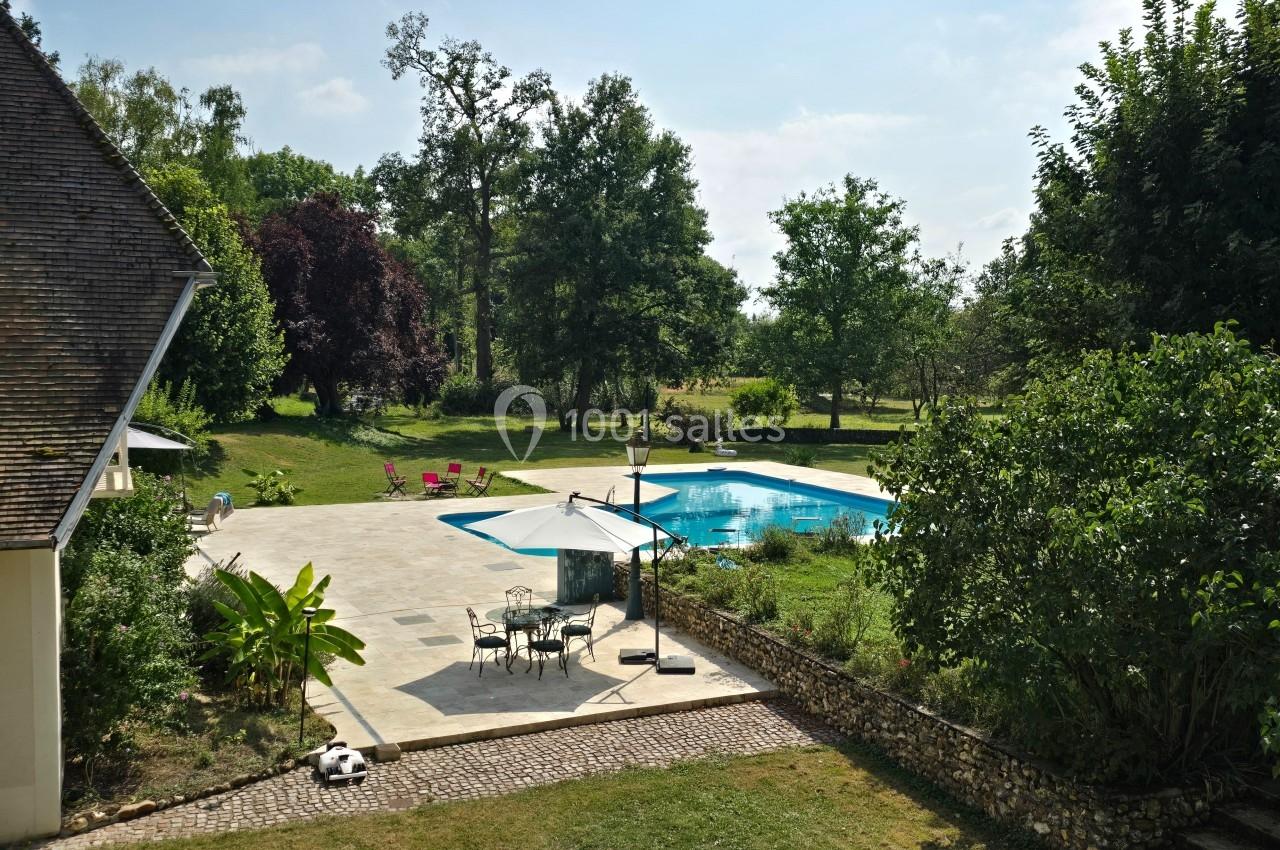 Terrasse avec parasol et mobilier de jardin près d'une piscine, entourée de verdure et d'arbres sous un ciel dégagé.