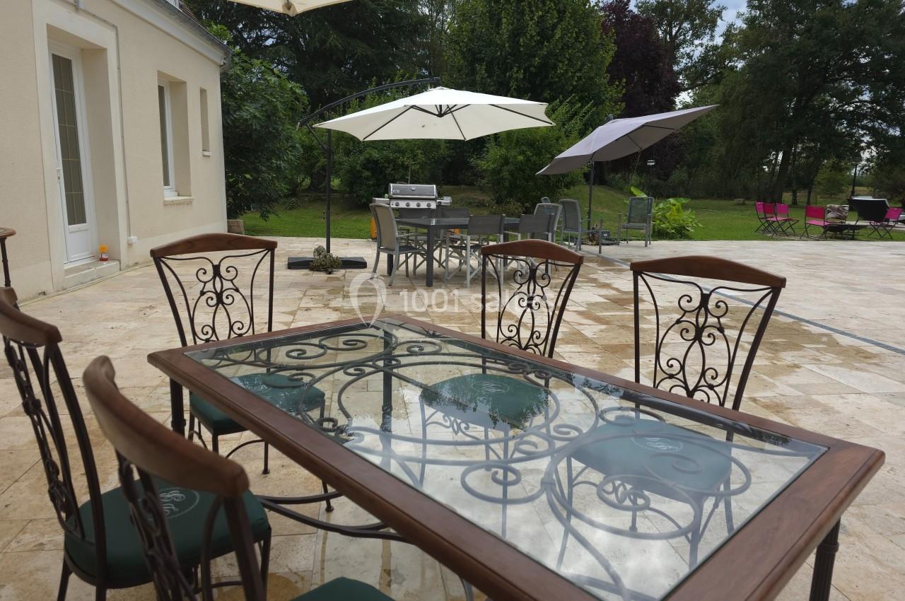 Table en verre avec chaises en métal sur une terrasse en pierre, entourée de parasols et d'un jardin verdoyant.