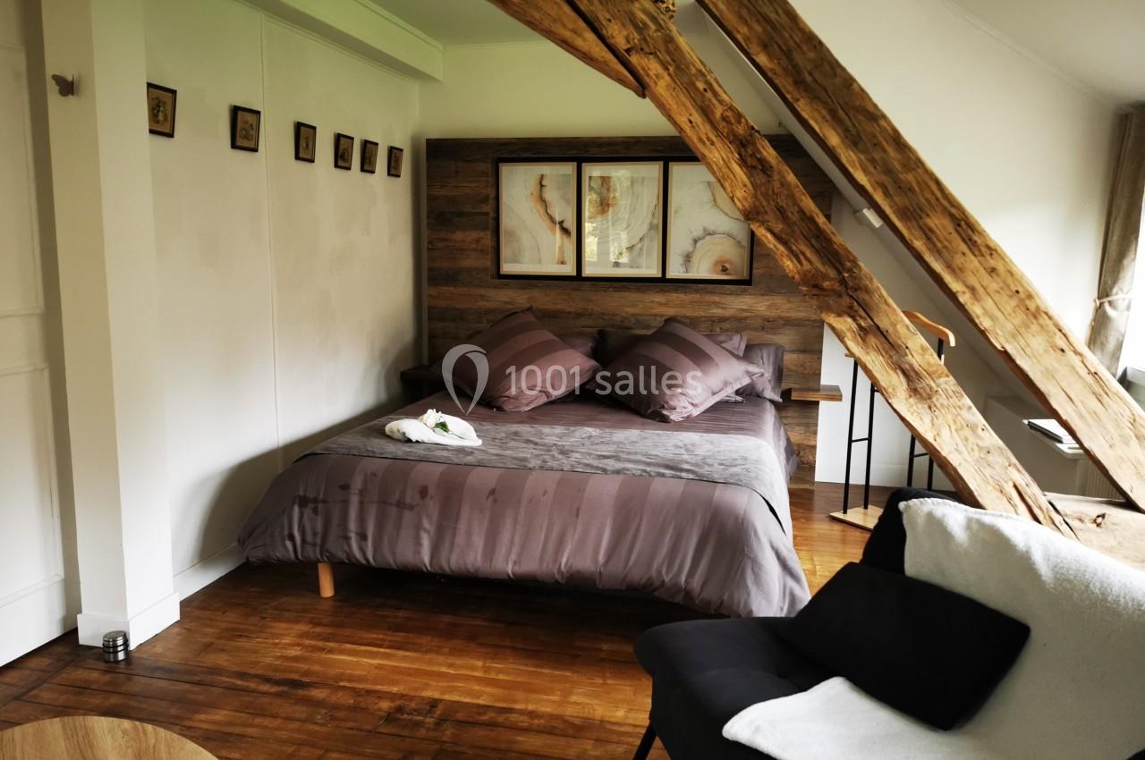 Chambre mansardée avec poutres apparentes, lit double aux draps gris, fauteuil noir et décoration murale en bois.