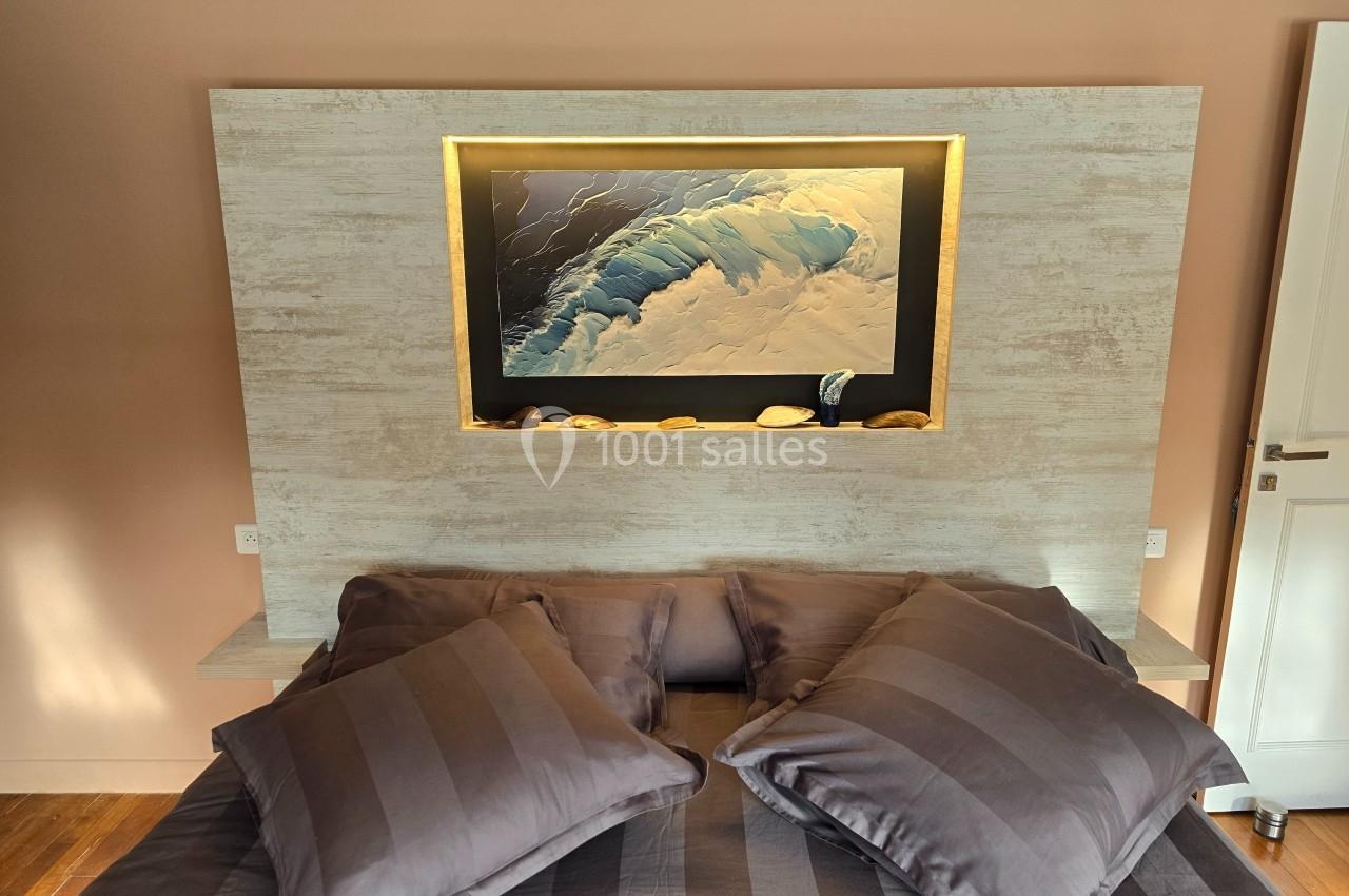 Lit avec coussins gris devant une tête de lit en bois clair, ornée d'un tableau de vague et d'une étagère éclairée.