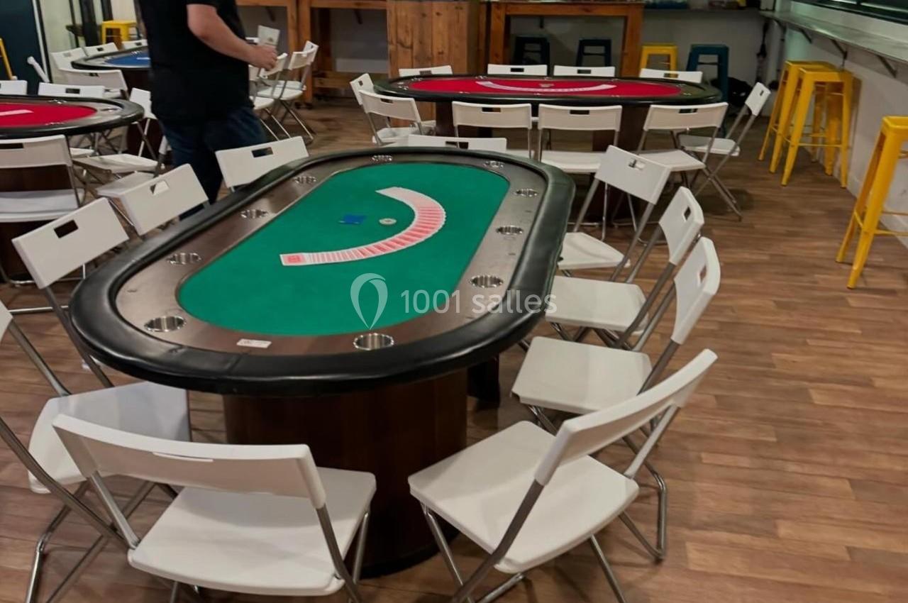 Salle de jeux avec tables de poker, chaises pliantes blanches et écran affichant des informations au mur.