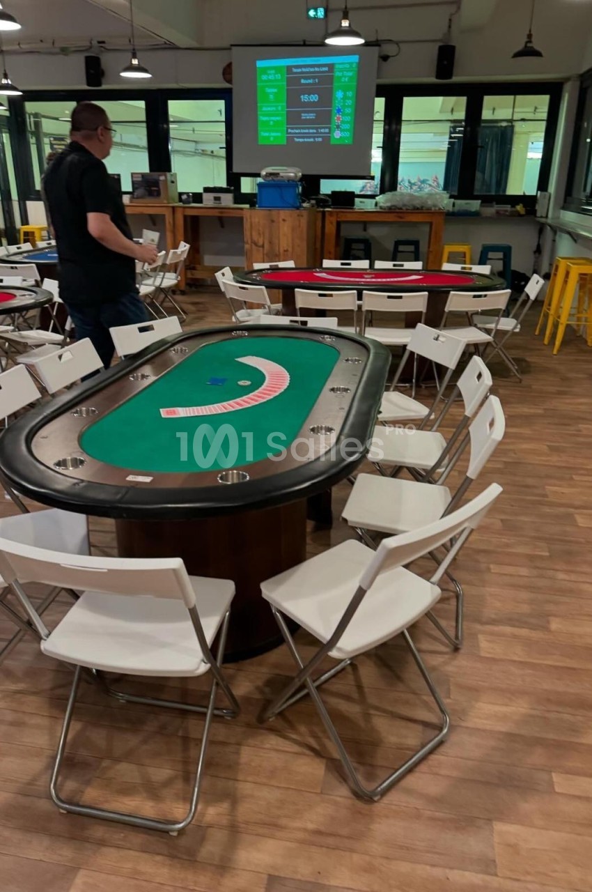 Salle de jeux avec tables de poker, chaises pliantes blanches et écran affichant des informations au mur.