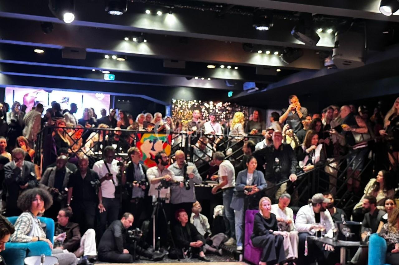 Salle bondée avec des personnes debout et assises, éclairage tamisé, ambiance animée lors d'un événement.
