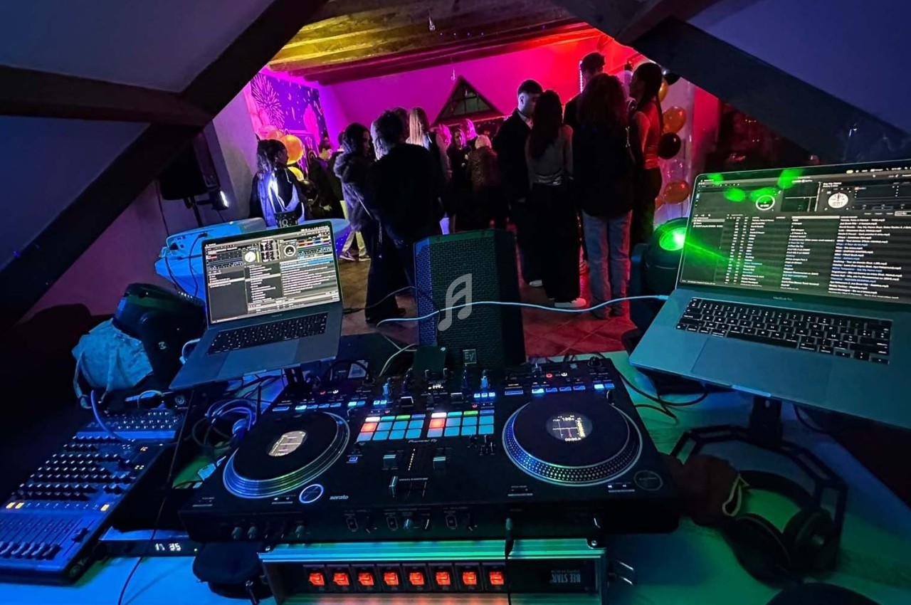 Vue depuis une table de mixage avec deux ordinateurs, face à une fête dans une pièce éclairée par des lumières colorées.