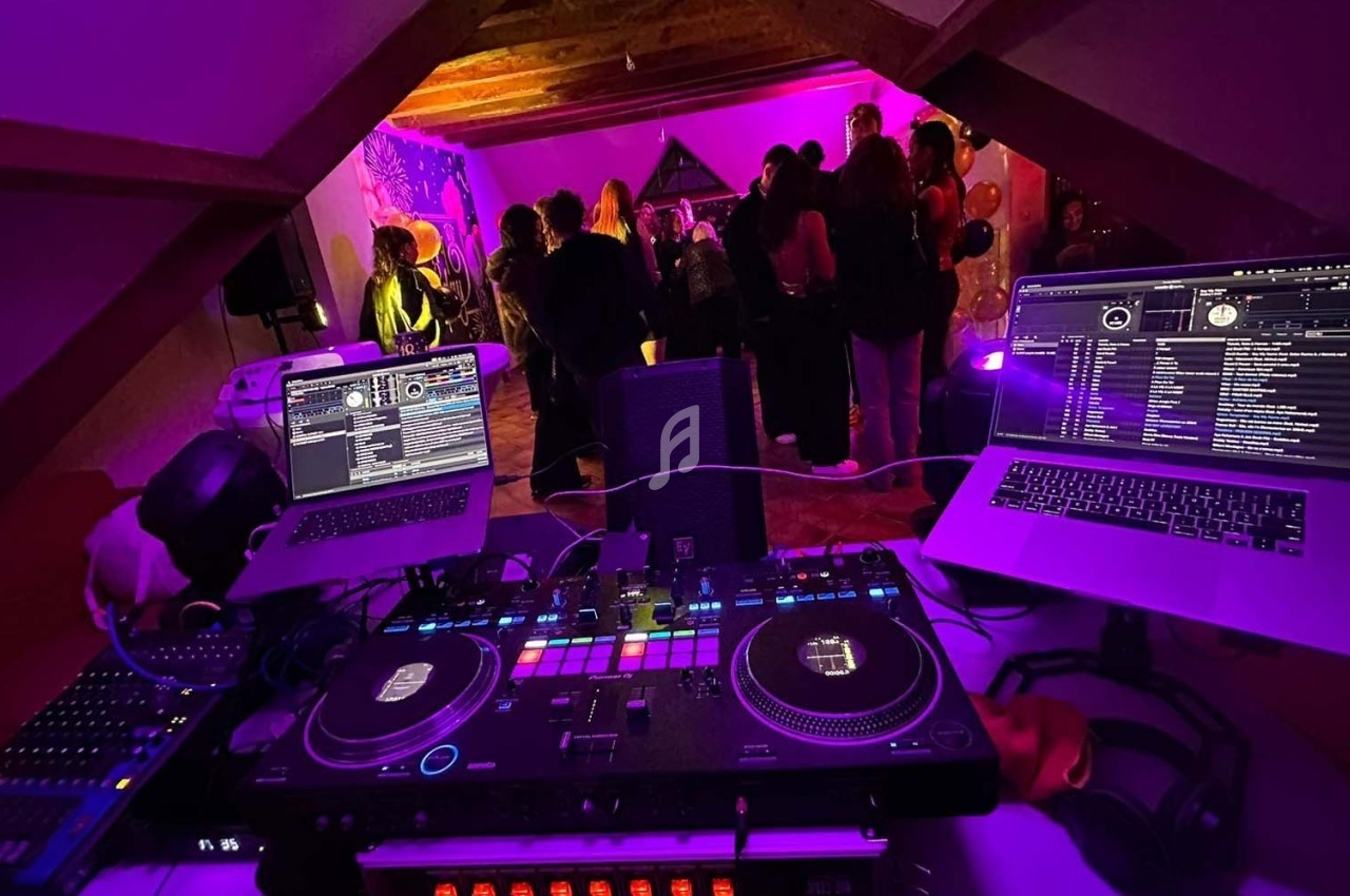 Vue depuis une cabine de DJ avec platines et ordinateurs, face à une piste de danse éclairée par des lumières colorées.