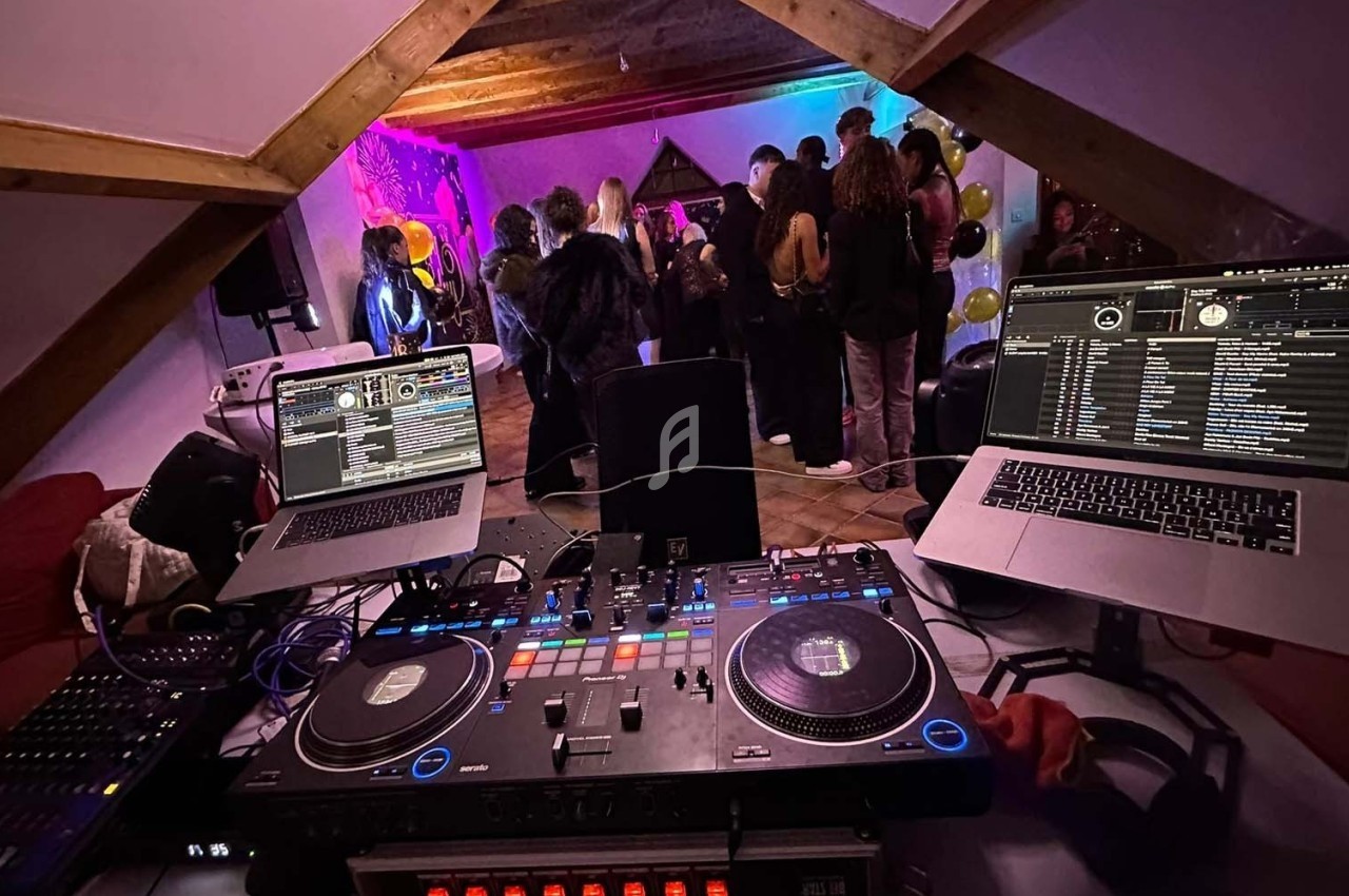Vue depuis une table de mixage avec ordinateurs, platines et lumières colorées, face à un groupe de personnes dansant.