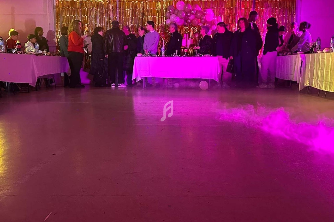 Salle décorée pour une fête avec des invités debout près de tables, éclairage coloré et fumée au sol.