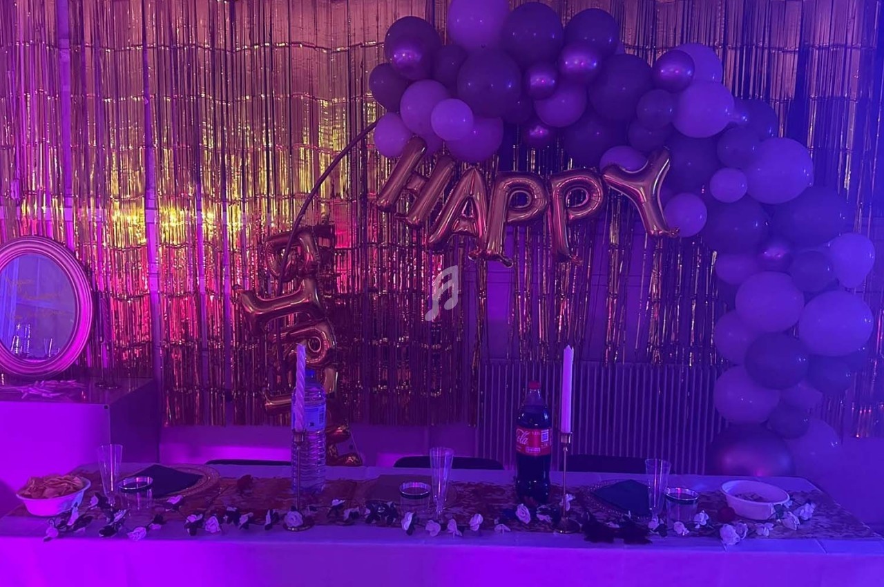 Décoration de fête avec ballons violets, guirlande ’Happy’, table dressée et fond doré scintillant.