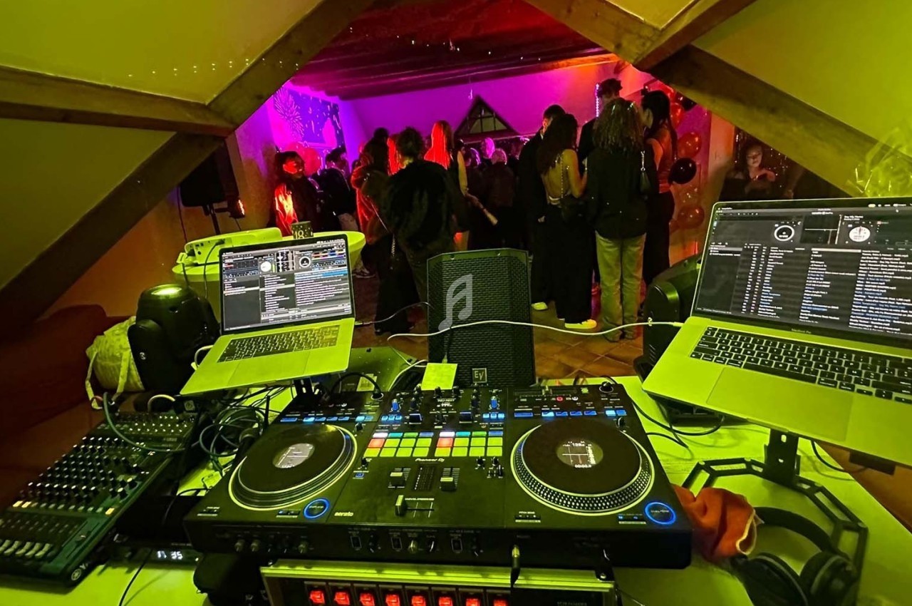Vue d'une table de mixage avec ordinateurs et platines, face à une salle où des personnes dansent sous des lumières colorées.
