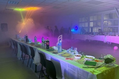 Salle obscure avec éclairage violet et vert, fumée diffuse, table dressée sur la gauche et rideaux brillants en arrière-plan.