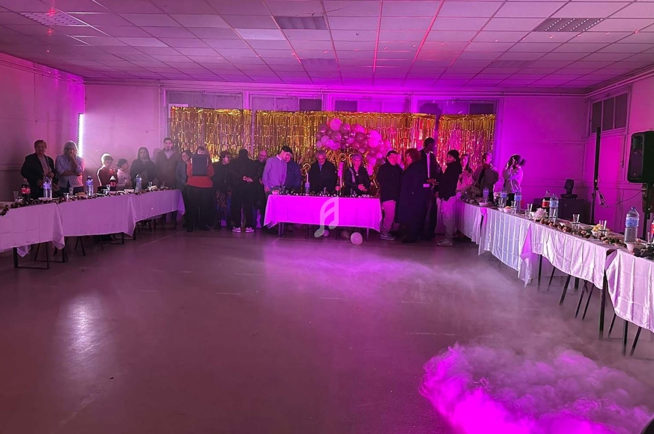 Salle décorée pour une fête avec des invités debout près d'une table, éclairage violet et fumée au sol.