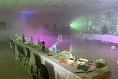 Salle obscure avec éclairage violet et vert, fumée diffuse, table dressée sur la gauche et rideaux brillants en arrière-plan.