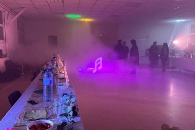 Salle obscure avec éclairage violet et vert, fumée diffuse, table dressée sur la gauche et rideaux brillants en arrière-plan.