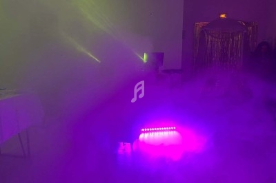 Salle obscure avec éclairage violet et vert, fumée diffuse, table dressée sur la gauche et rideaux brillants en arrière-plan.