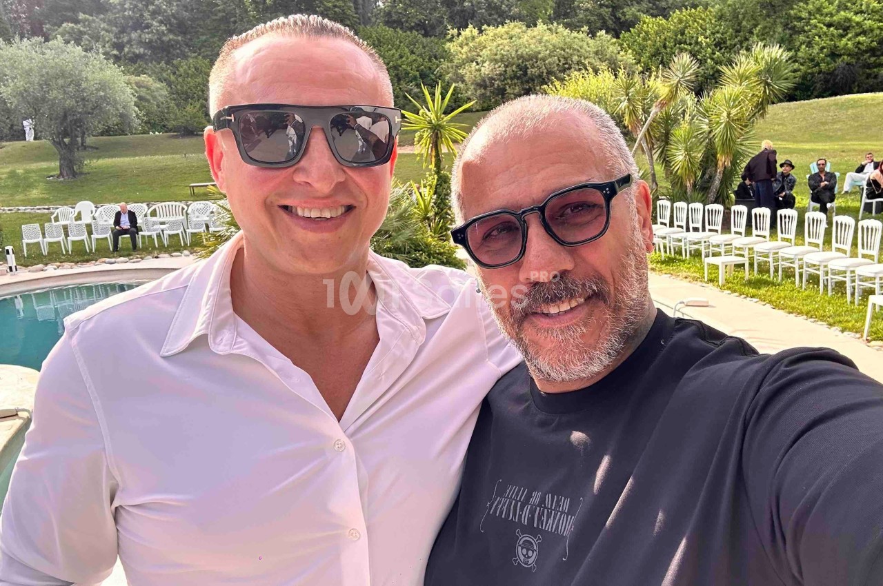 Deux hommes souriants posent pour un selfie près d'une piscine, avec des chaises blanches et un jardin en arrière-plan.