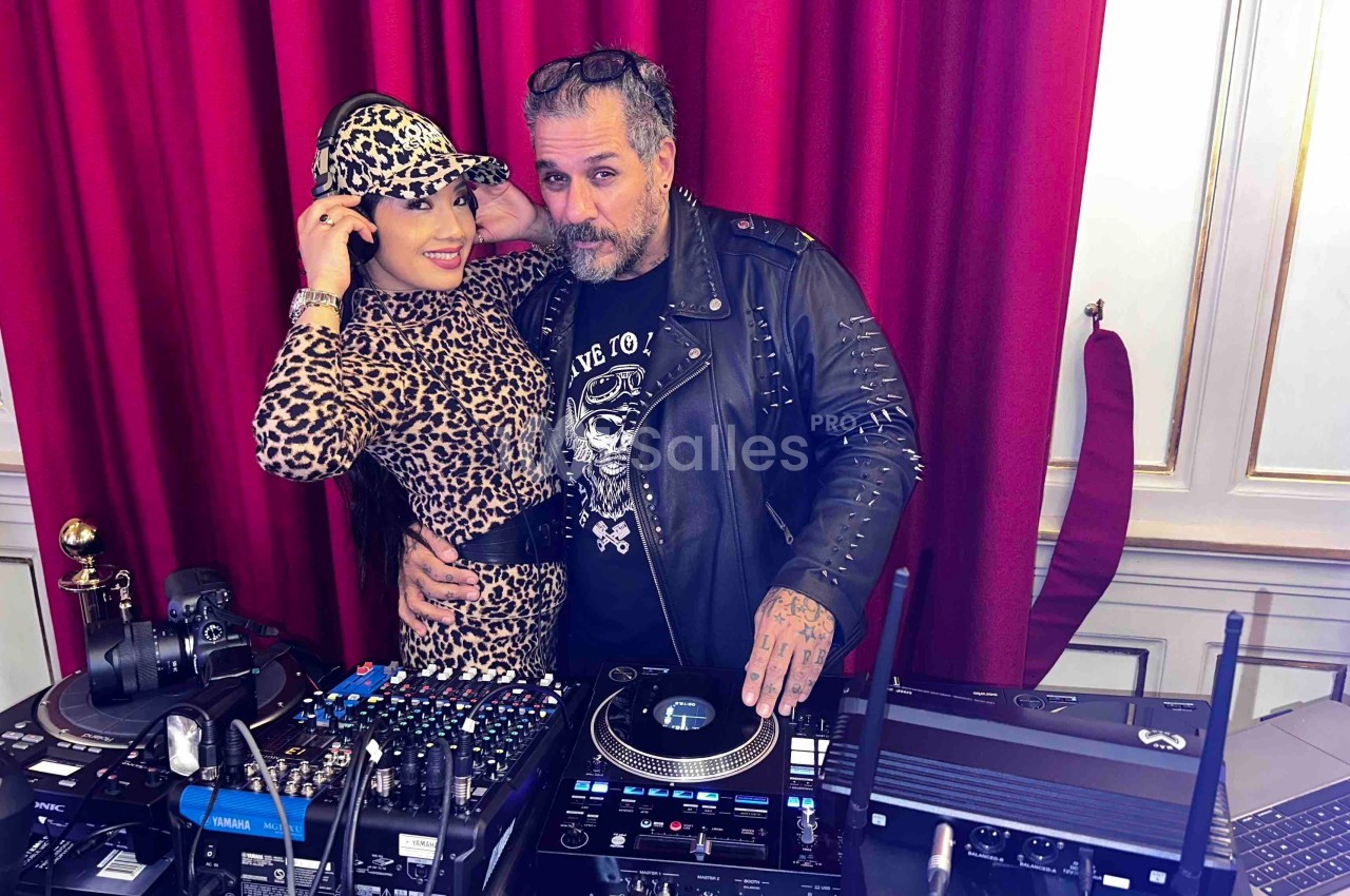Une femme en tenue léopard et un homme en veste en cuir posent derrière une table de mixage avec des platines DJ.