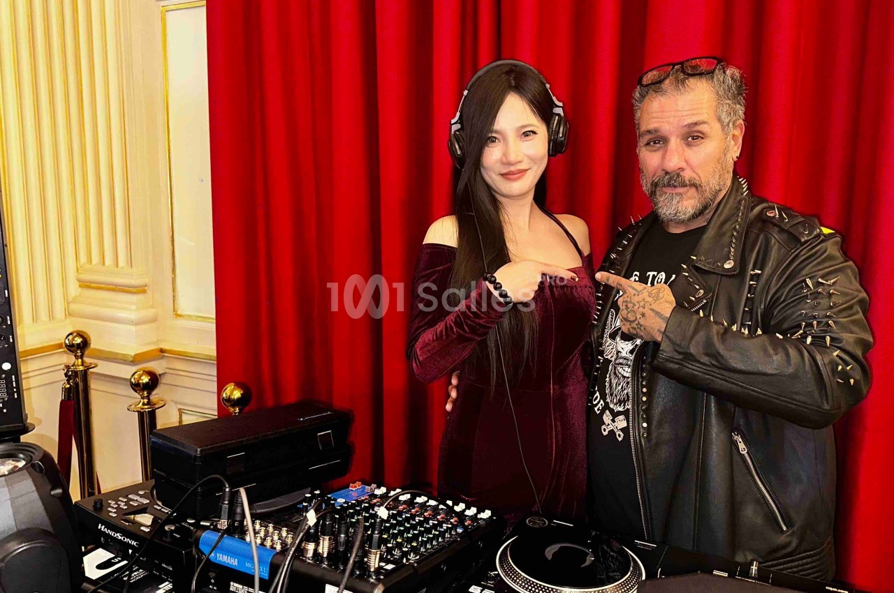 Une femme et un homme posent devant une table de mixage, avec un rideau rouge en arrière-plan.