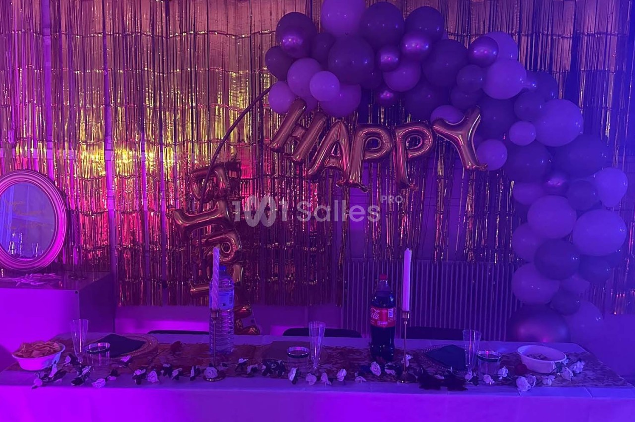 Décoration festive avec ballons violets et dorés, guirlande ’Happy’, table dressée et fond doré scintillant.