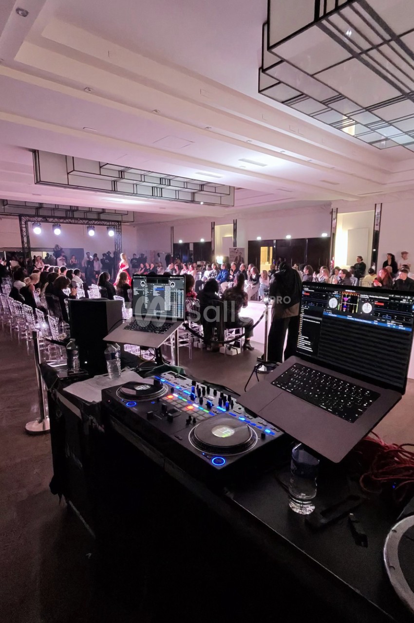 Table de mixage DJ et ordinateurs portables au premier plan, face à une salle où des invités assis sur des chaises transparentes suivent un événement sous un éclairage rosé.