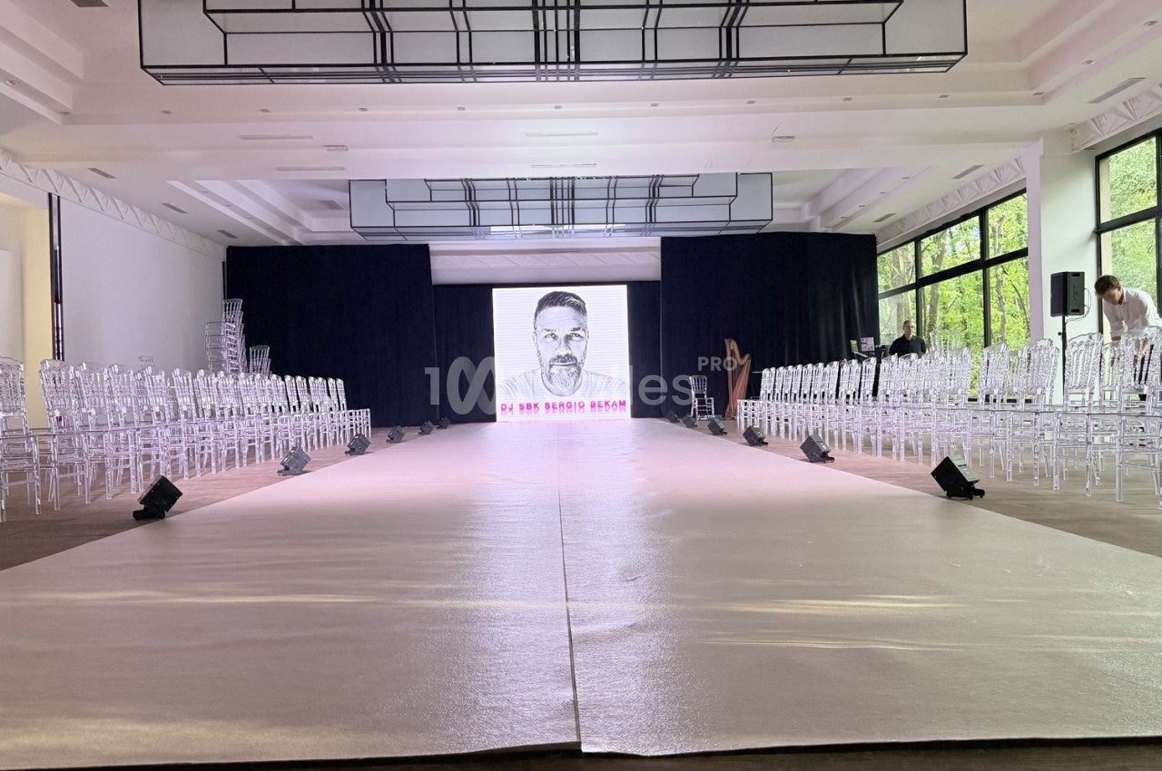 Podium de défilé à tapis argenté, bordé de chaises transparentes, devant un écran LED dans une salle lumineuse.