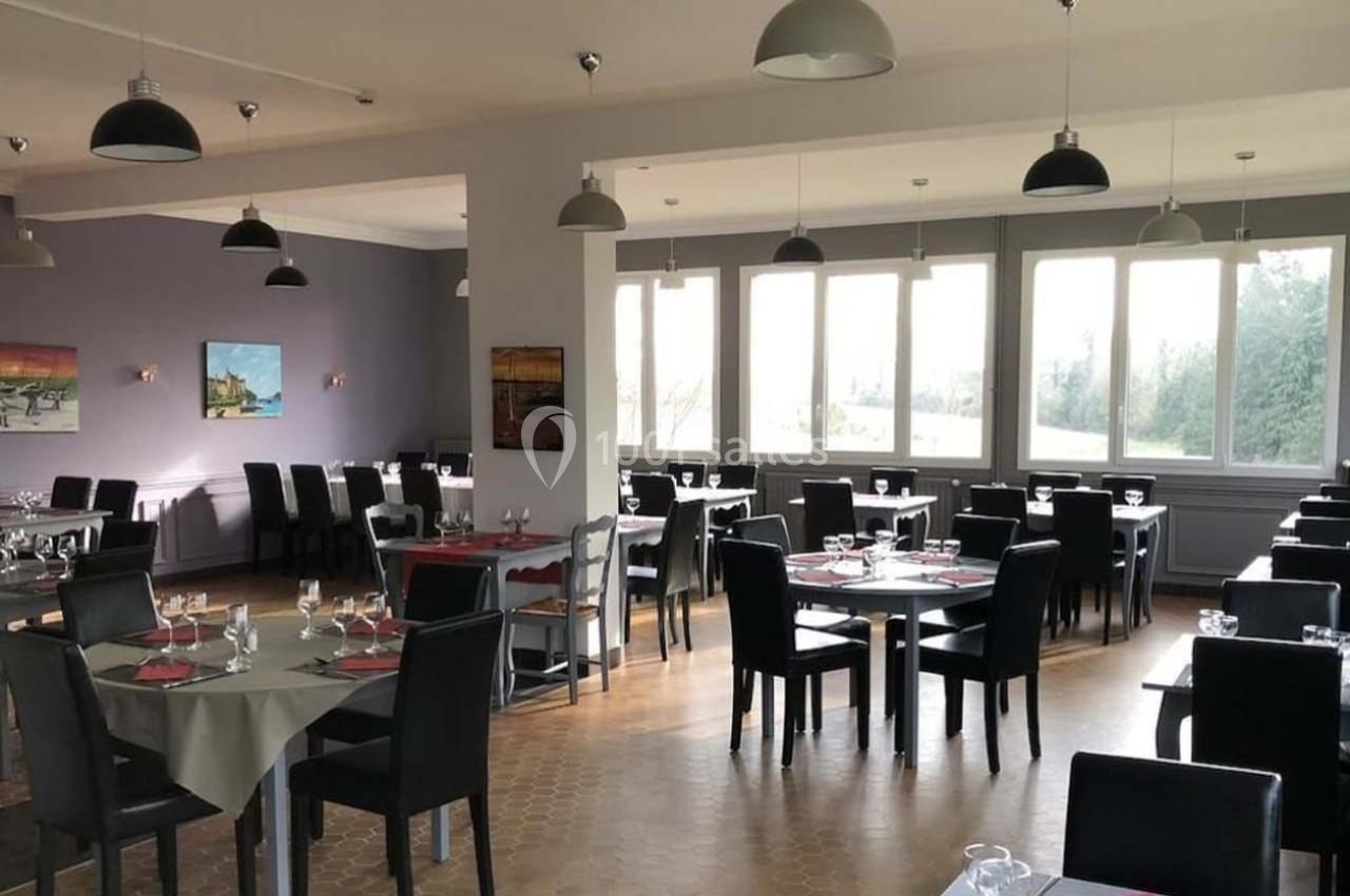 Salle de restaurant lumineuse avec grandes fenêtres, tables dressées et chaises noires, décorée de tableaux colorés.