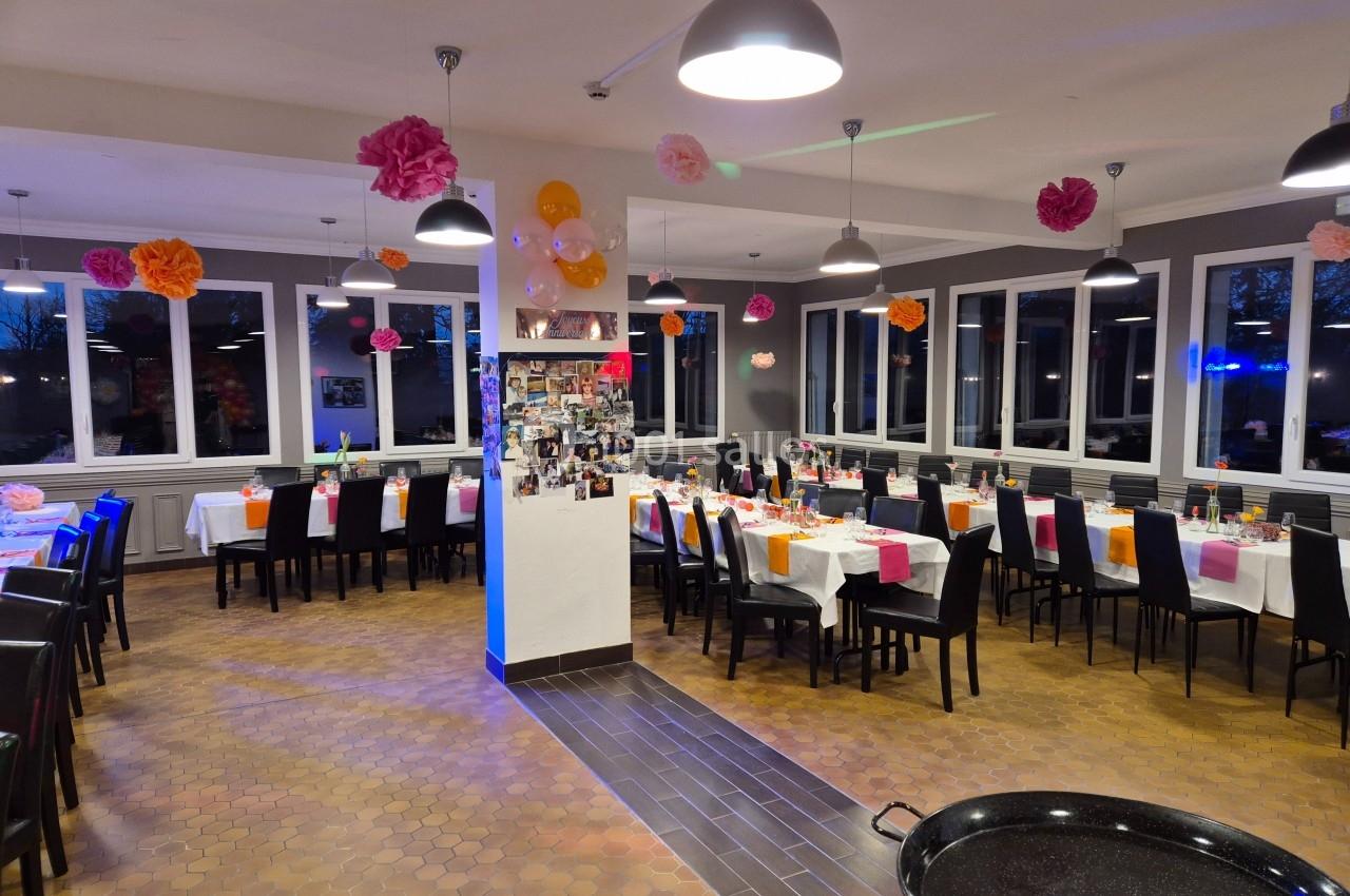 Salle de réception décorée avec des tables dressées, des chaises noires et des guirlandes de fleurs colorées suspendues.