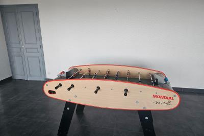 Miniature Location salle Le Teilleul (Manche) - Le Grand Gîte du Lac #16 Baby-foot en bois clair avec pieds noirs, situé dans une pièce au sol sombre et murs clairs avec une porte grise.