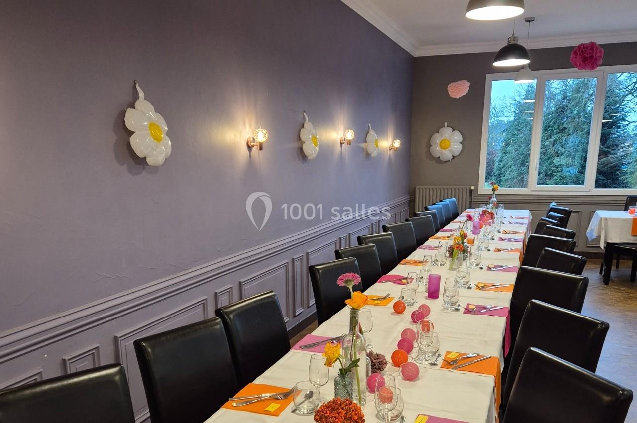 Salle décorée avec des tables alignées, nappes blanches, chaises noires et décorations florales sur un mur violet.
