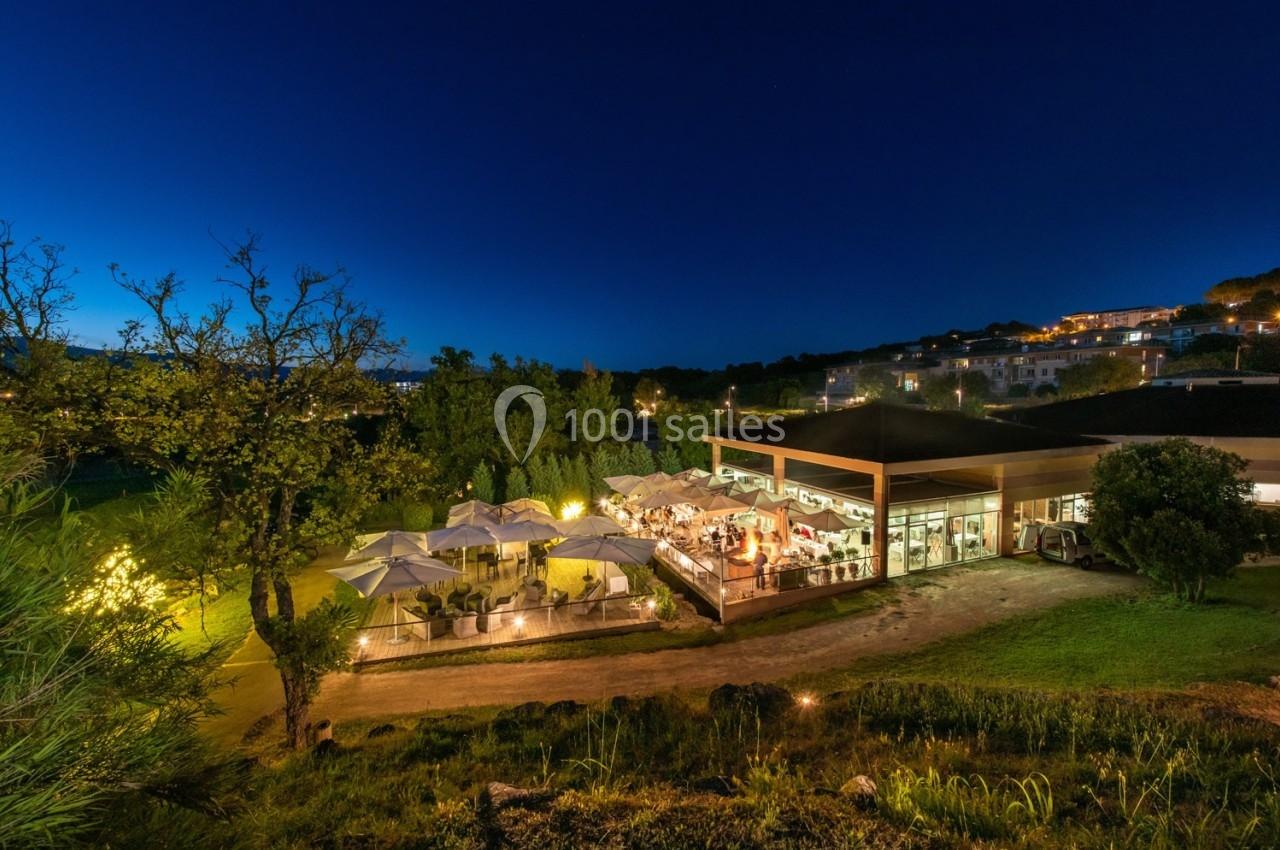Terrasse d'un restaurant éclairée en soirée, entourée de végétation et située près d'un bâtiment moderne.