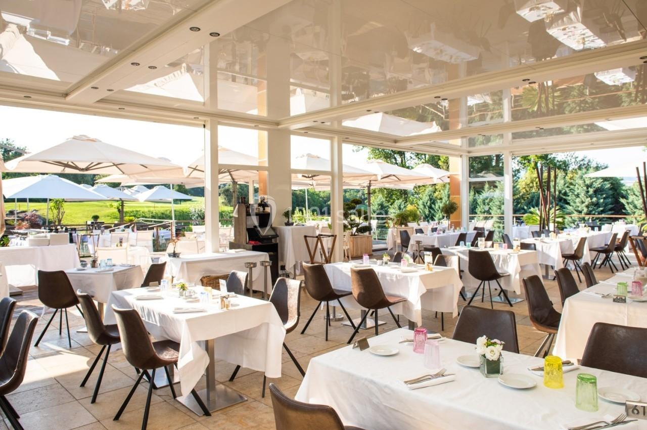 Salle de restaurant lumineuse avec grandes baies vitrées, tables dressées et terrasse extérieure avec parasols.