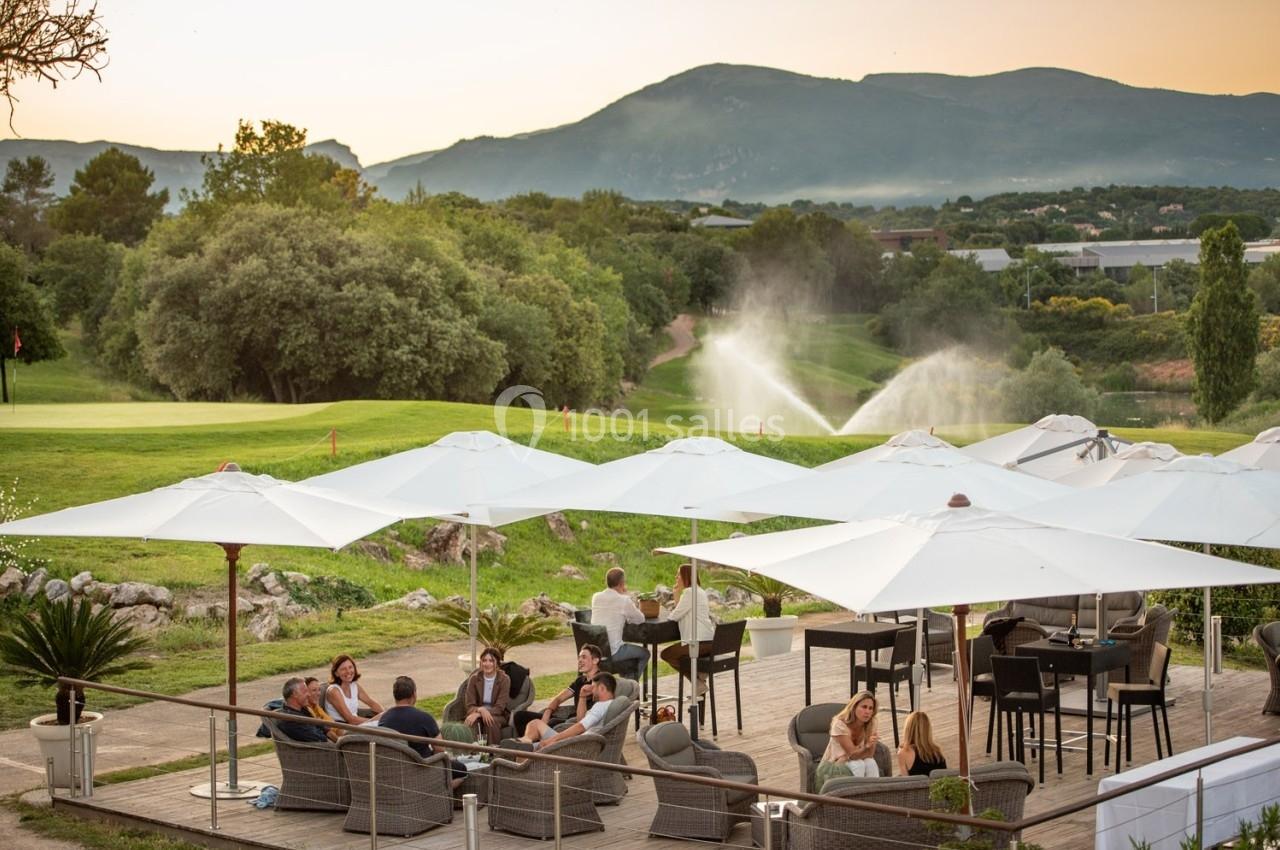 Terrasse avec des tables et des parasols, des personnes assises, vue sur un terrain de golf et des montagnes en arrière-plan.